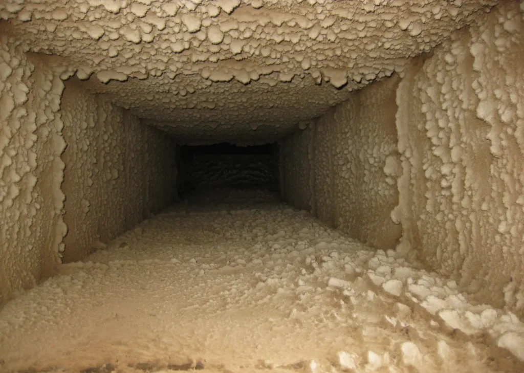 A rectangular air duct with walls and ceiling covered in dust and debris, viewed from the inside. The grime and buildup create an uneven, textured surface throughout the duct's interior, indicating it hasn't been cleaned in a long time. It's evident this space is overdue for professional air duct cleaning.