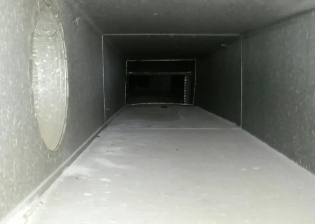 A view inside a rectangular ventilation duct with a side circular opening reveals the need for air duct cleaning. The duct appears dusty and grimy, with a buildup of dirt visible along the edges and floor. The lighting is dim, casting shadows along the interior surfaces.