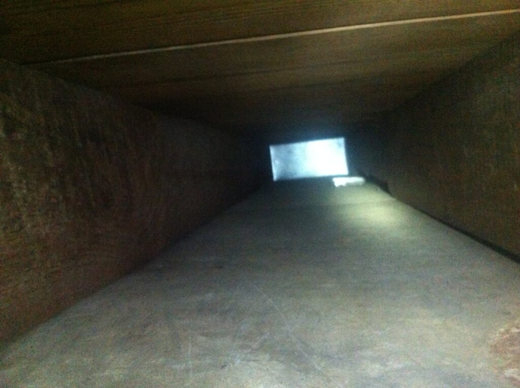 A view inside a long, rectangular ventilation duct with a metallic interior. Light is visible at the far end, indicating an opening or exit. The surface is smooth with minimal debris thanks to recent air duct cleaning, and the edges are straight and parallel.