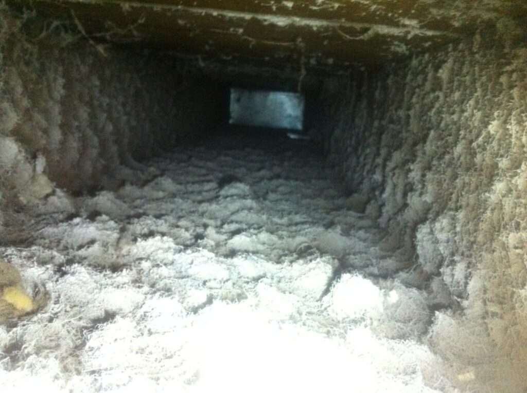 A close-up view of the inside of a duct reveals a significant buildup of dust and debris along the walls and floor. The accumulation has formed thick, fluffy layers, underscoring the urgent need for air duct cleaning.
