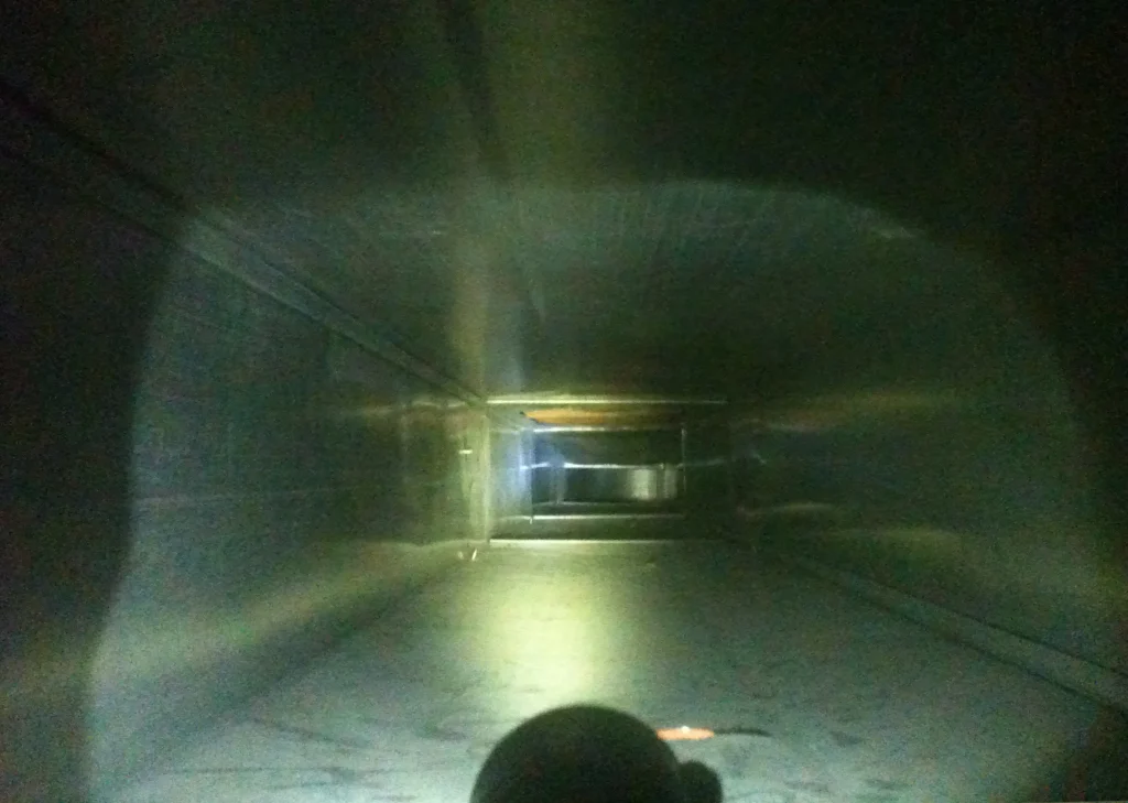 A narrow, rectangular air duct is shown from an inside perspective, illuminated by a flashlight. The light reveals the metallic walls and ceiling extending into the distance, with some reflections visible. The image has a dark and enclosed atmosphere, highlighting the need for air duct cleaning.